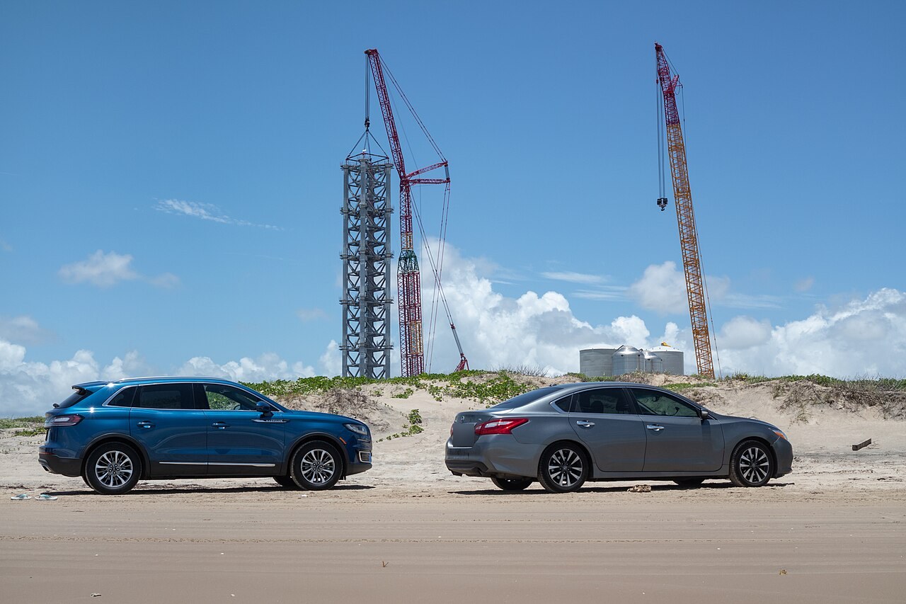 Two cars parked on Boca Chica Beach in Texas, with SpaceX's Starship launch tower under construction and cranes rising just behind the dunes
