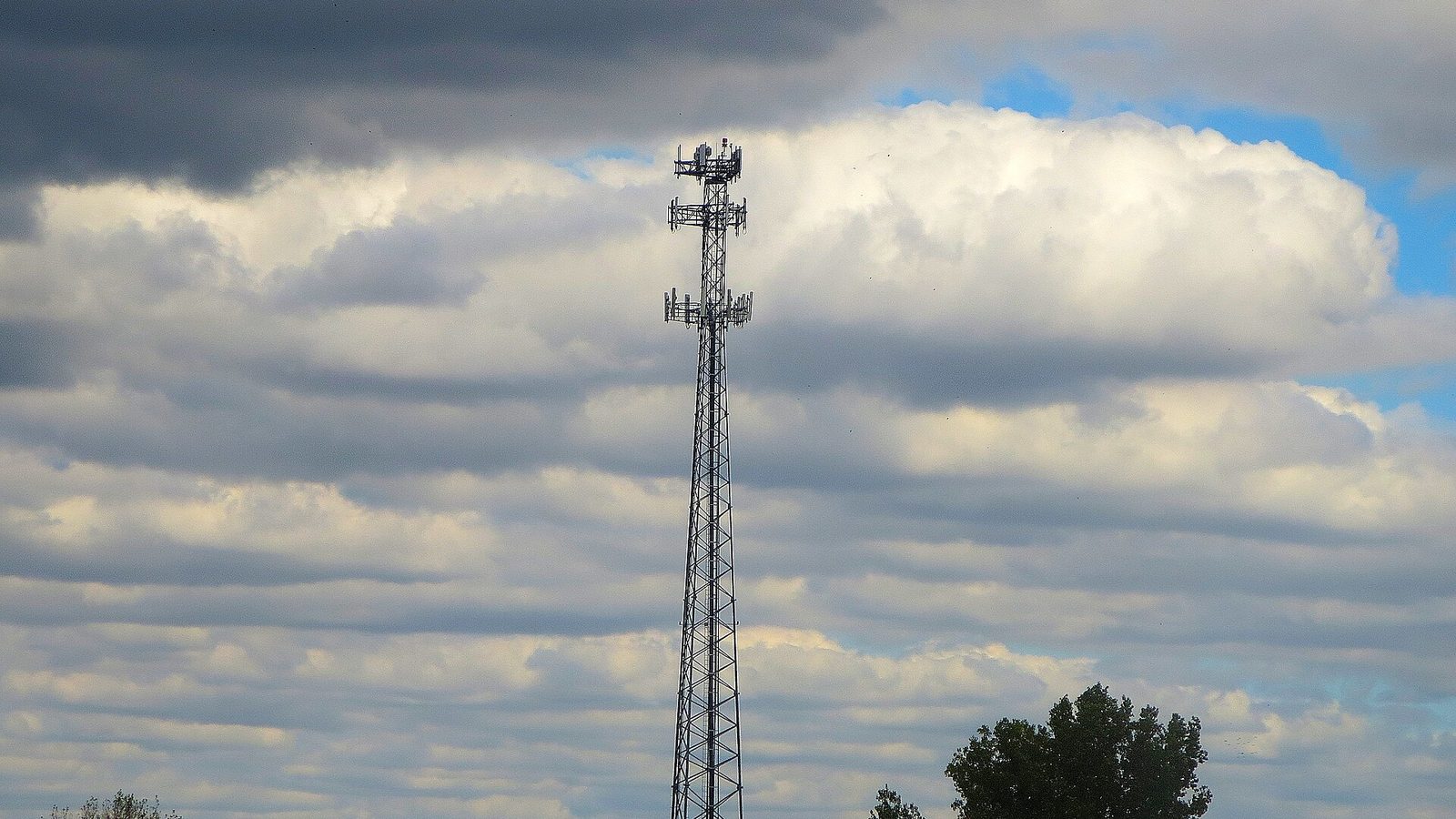 A tall steel lattice cell tower with antennas at the top stands over a harvested field and a line of trees under a cloudy sky
