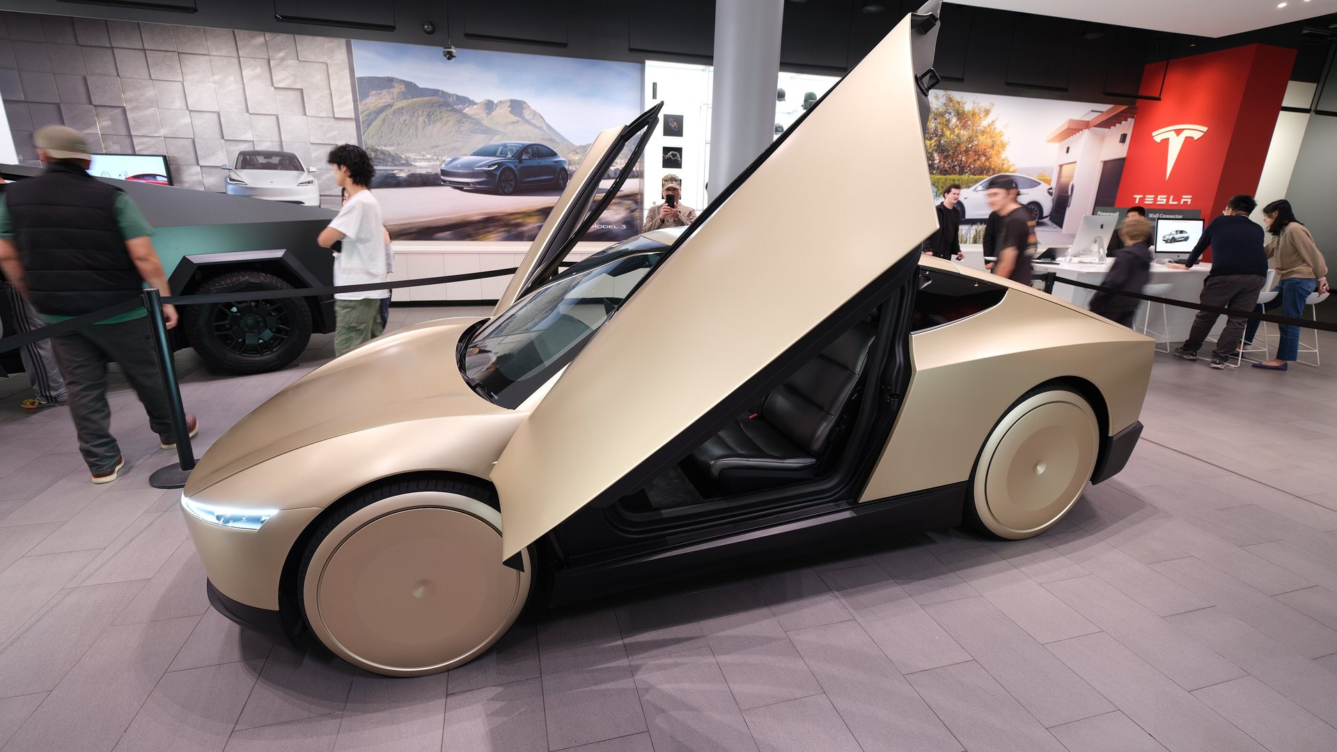 A champagne-gold, two-door Tesla Cybercab with its butterfly doors raised, on display inside a Tesla showroom with visitors and a Cybertruck in the background