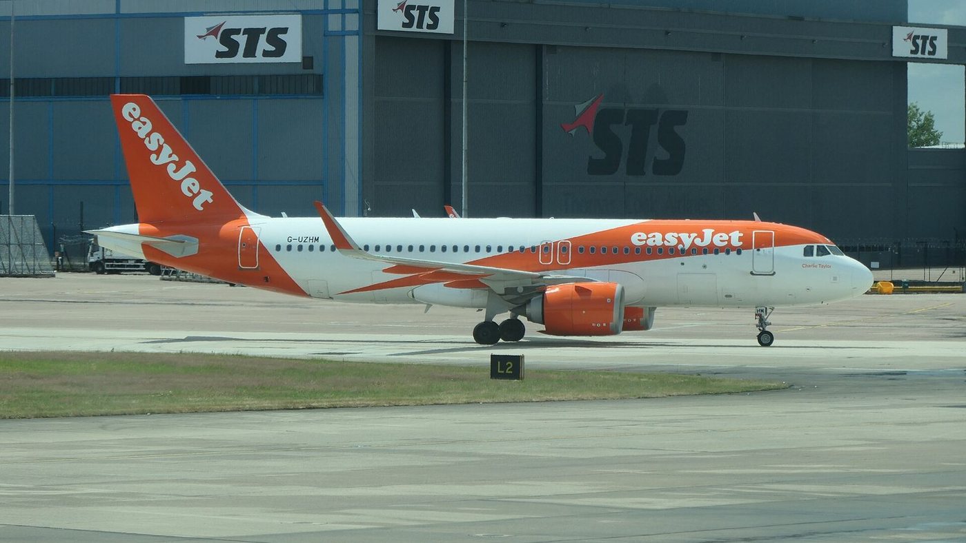 An orange and white easyJet Airbus A320neo, registration G-UZHM, taxiing past a hangar