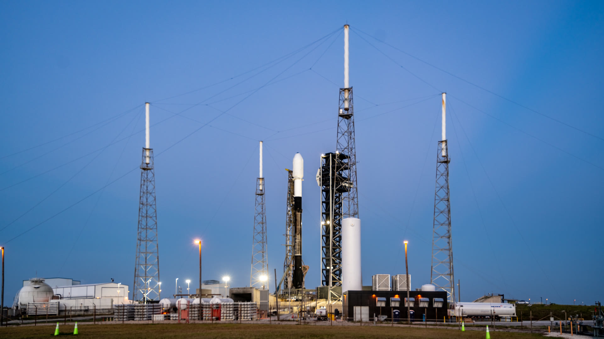 A SpaceX Falcon 9 rocket standing on the launch pad at Space Launch Complex 40 at dawn, framed by four tall lightning-protection towers under a blue sky
