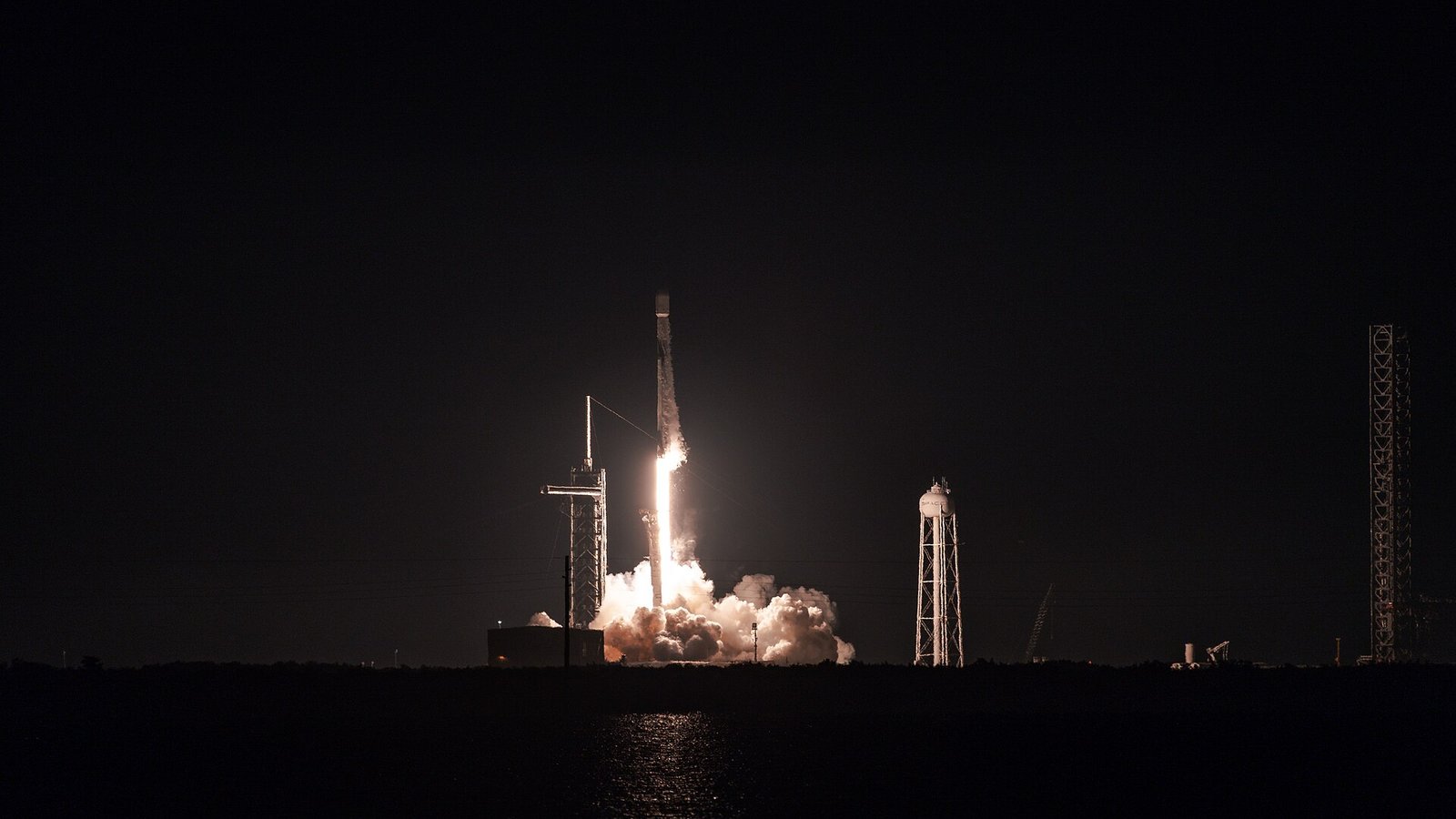 A SpaceX Falcon 9 rocket lifts off at night from Launch Complex 39A at Kennedy Space Center, lighting up a cloud of exhaust beside the launch tower