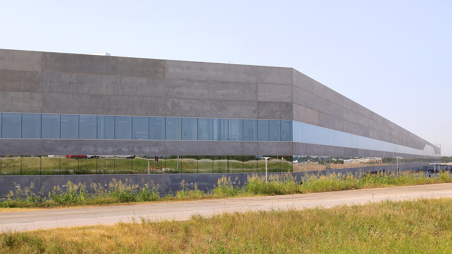 The long grey concrete and glass facade of Tesla's Gigafactory Texas Building 1 near Austin, seen across a grassy roadside on a clear day