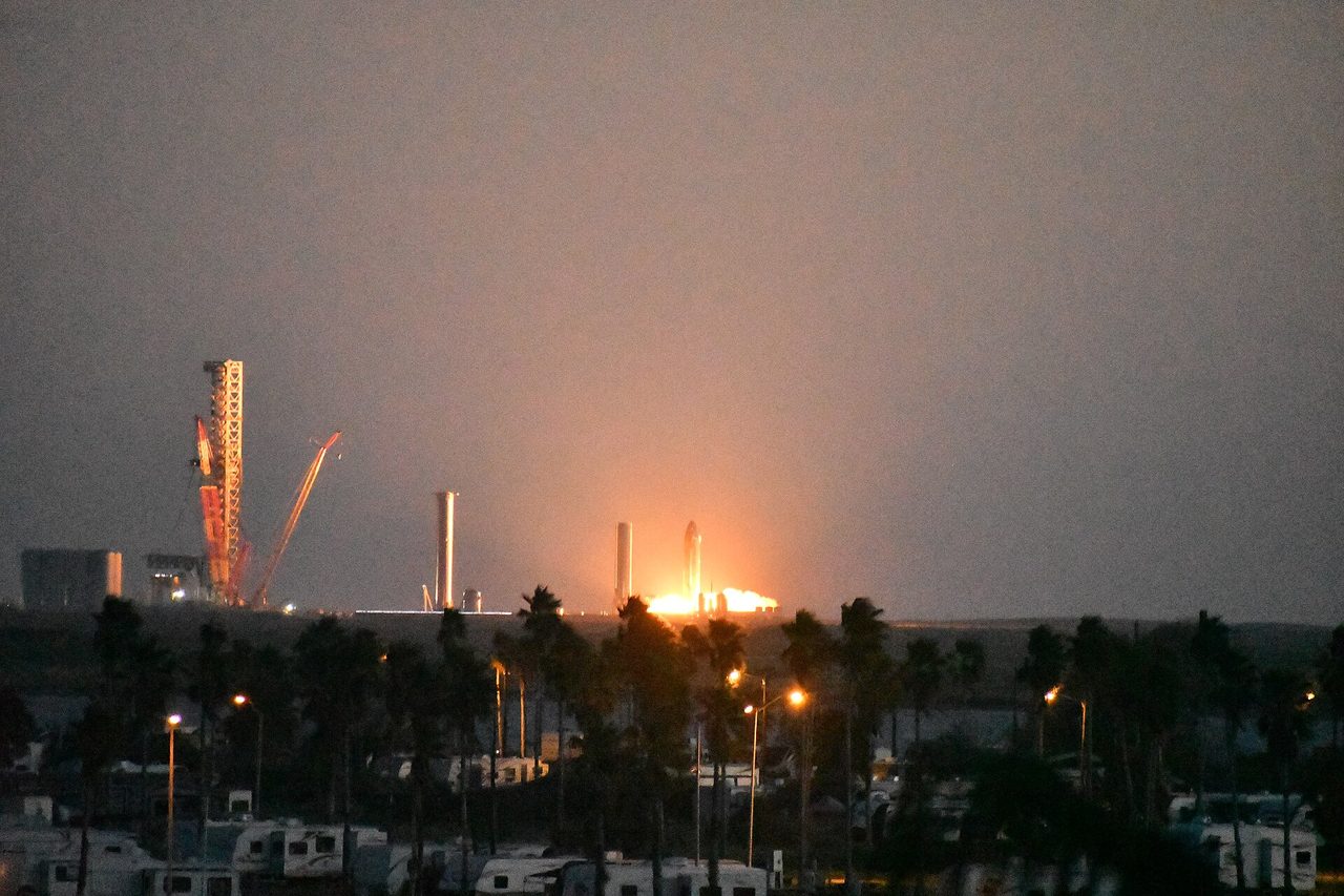 An earlier Starship static fire lights up the Starbase skyline at dusk, seen across palm trees from South Padre Island
