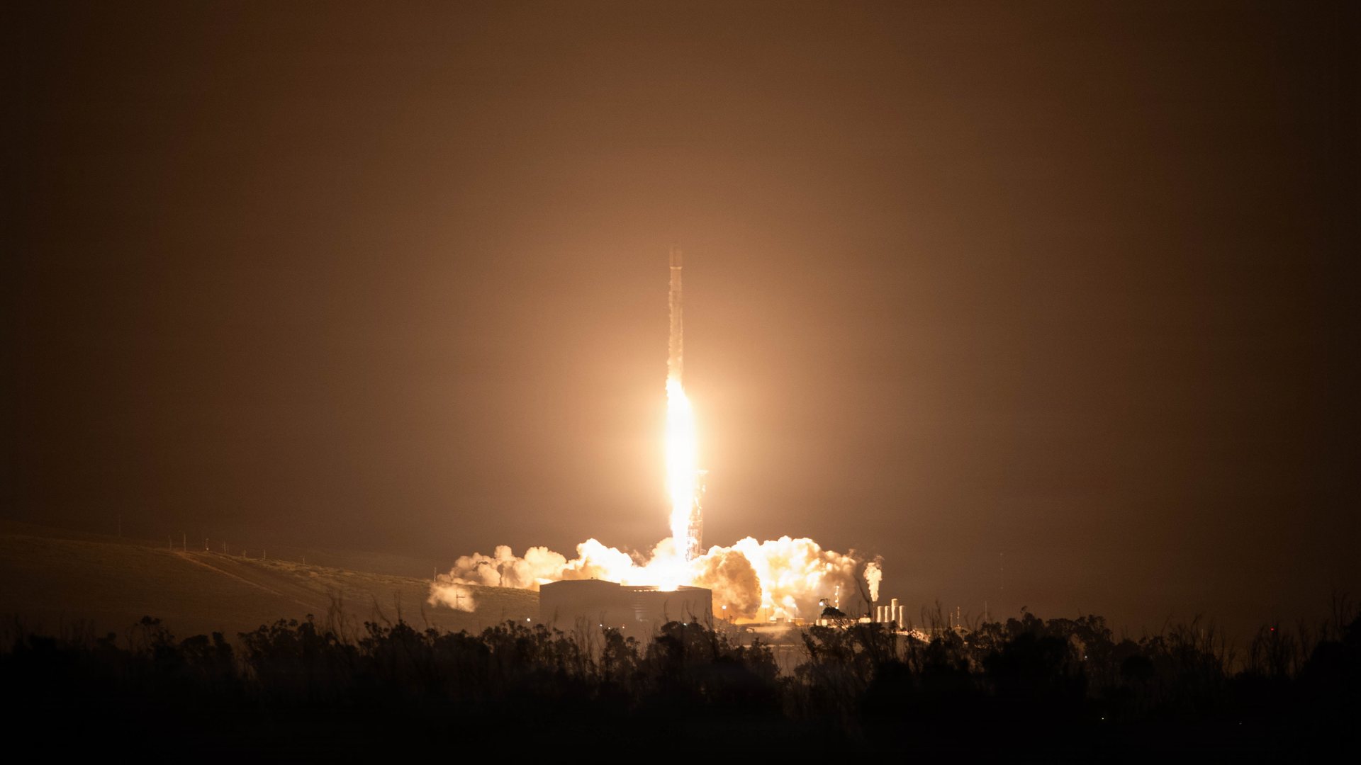 A Falcon 9 rocket lifts off at night from Vandenberg, its exhaust lighting up the clouds of steam around the pad