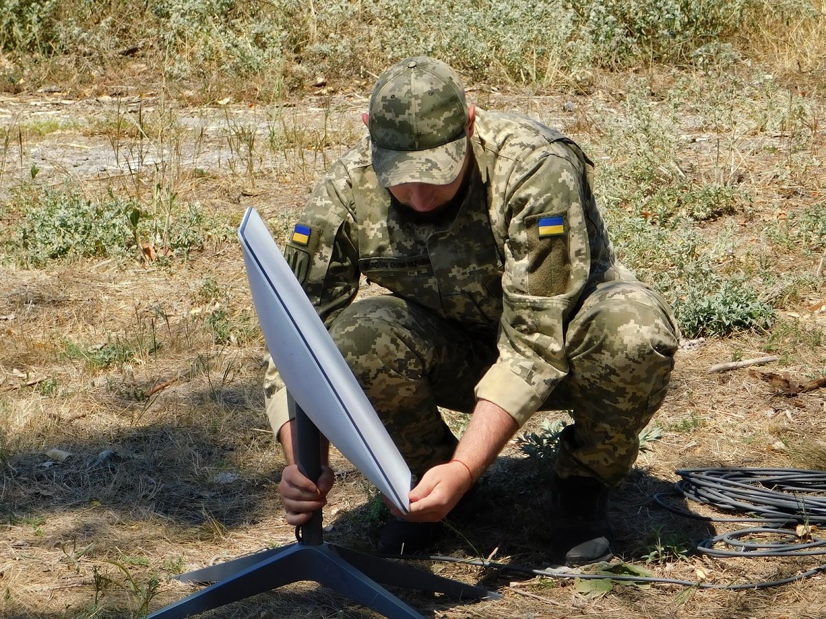 A Ukrainian soldier in camouflage crouches on dry grass, setting up a white rectangular Starlink dish on its stand