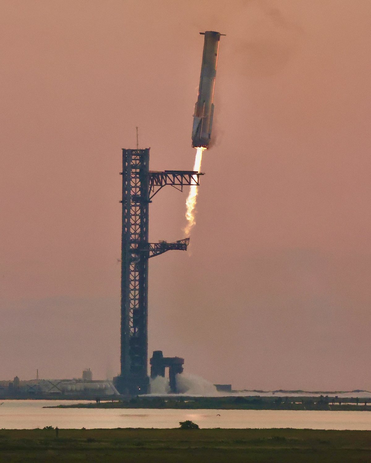 A Super Heavy booster, engines firing, descends at an angle toward the tall steel launch tower at Starbase, whose two catch arms are open, against a pink dawn sky