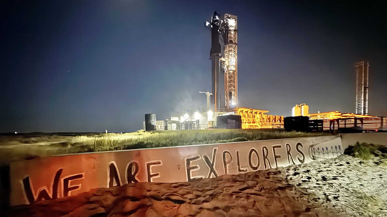 A fully stacked SpaceX Starship stands lit up beside its launch tower at night at Starbase, Texas, behind a beach barrier painted with the words We Are Explorers