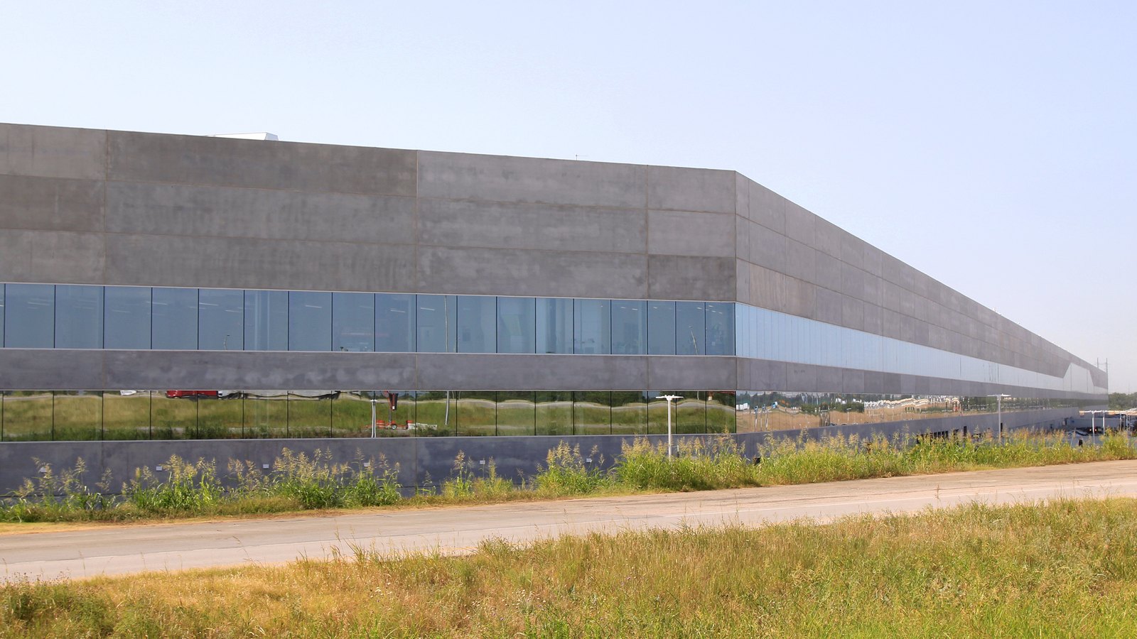 The long grey concrete and glass front of Tesla Gigafactory Texas Building 1 behind a road and tall grass