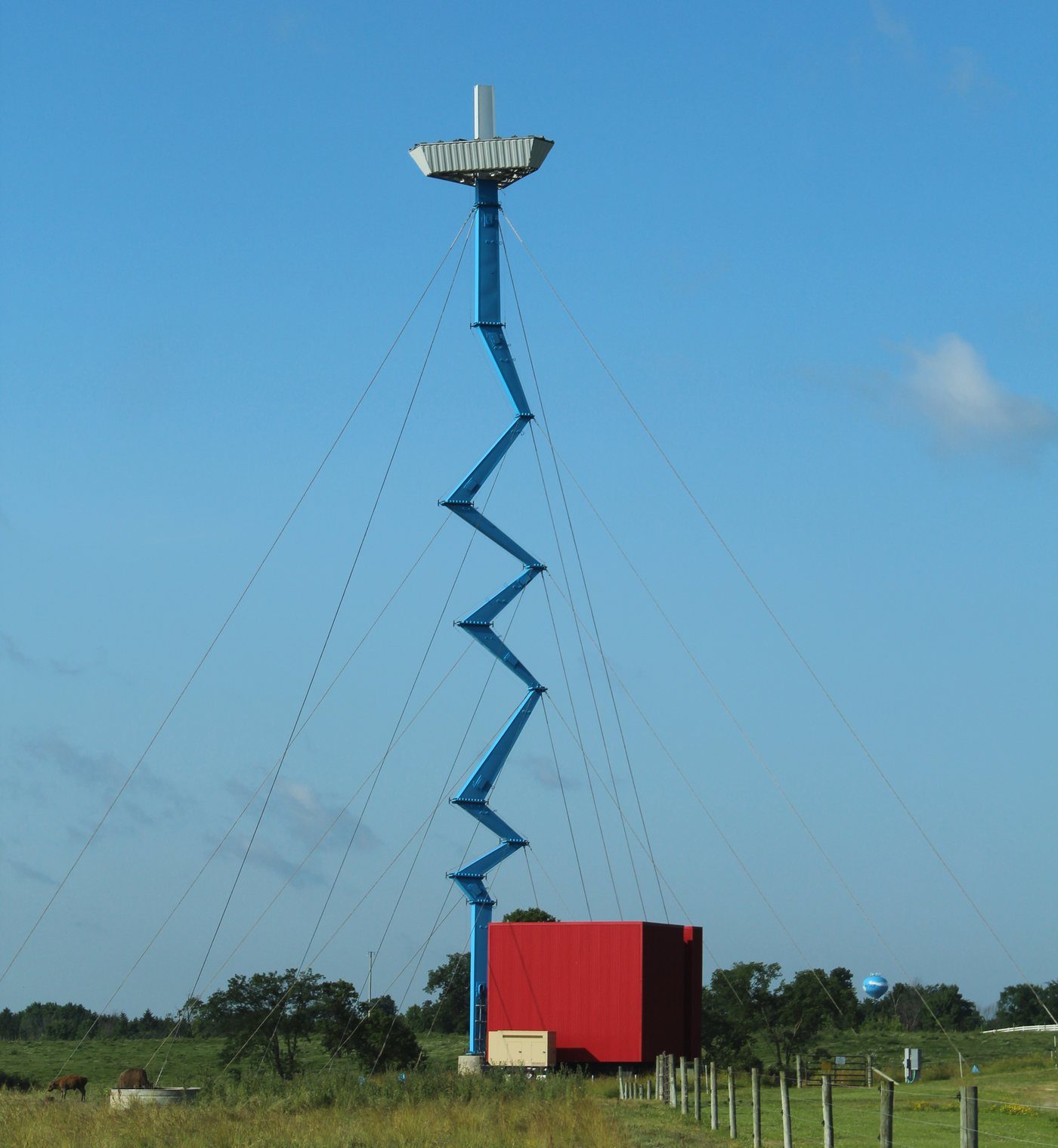 A blue Verizon cell tower with a zigzag design above a red equipment shelter in a field